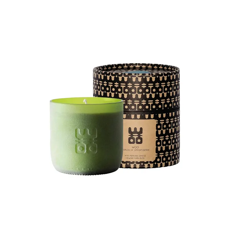 Groene kaars met wit lontje en WOO-logo naast een zwart-beige cilindrische doos met patroon en WOO-etiket.